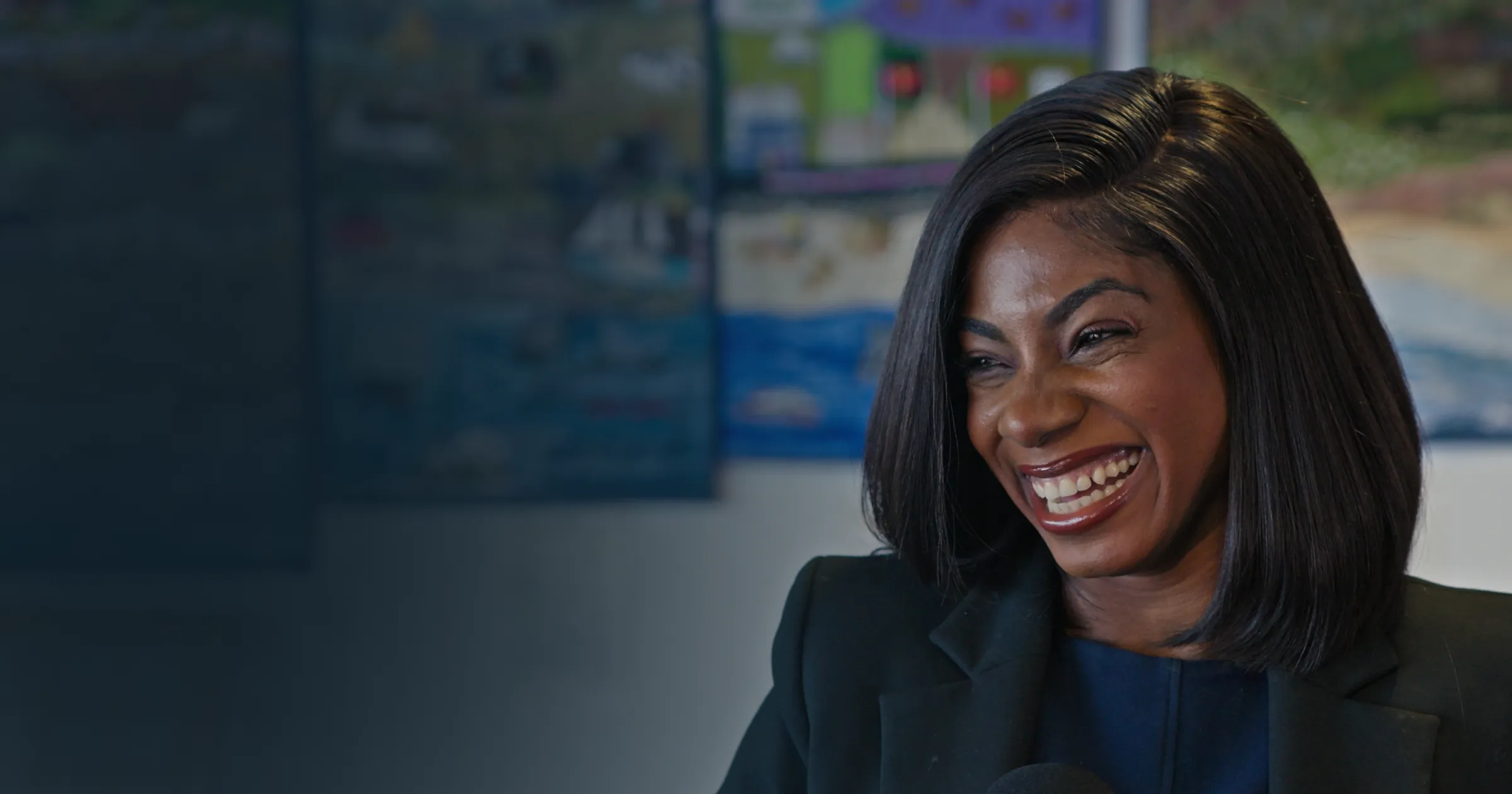 Smiling woman with dark hair and a blazer stands in front of a blurred classroom wall display, conveying a sense of joy and confidence.